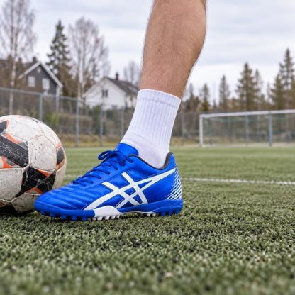 Blå fotbollssko med snörning, vit sula och mönster, sedd från sidan på gräs.